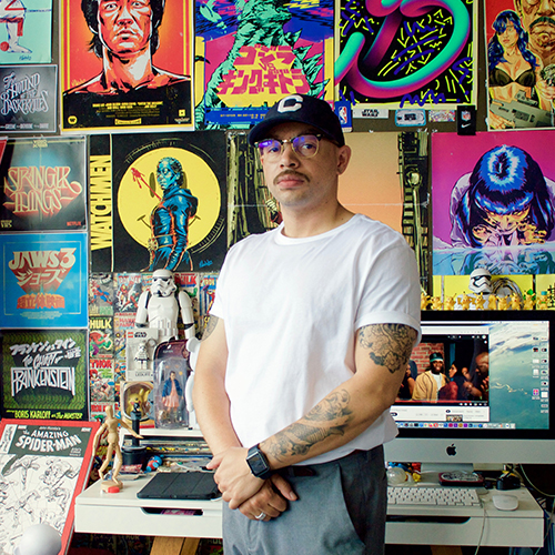 Photograph of a man in a white t shirt, black cap, and glasses with his hands crossed. He has a mustache and tattoos on his arms. A colorful wall of graphics can be seen behind him.