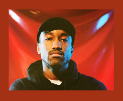 Photograph of Richard A Chance in a black hat and black sweatshirt in front a a red backdrop