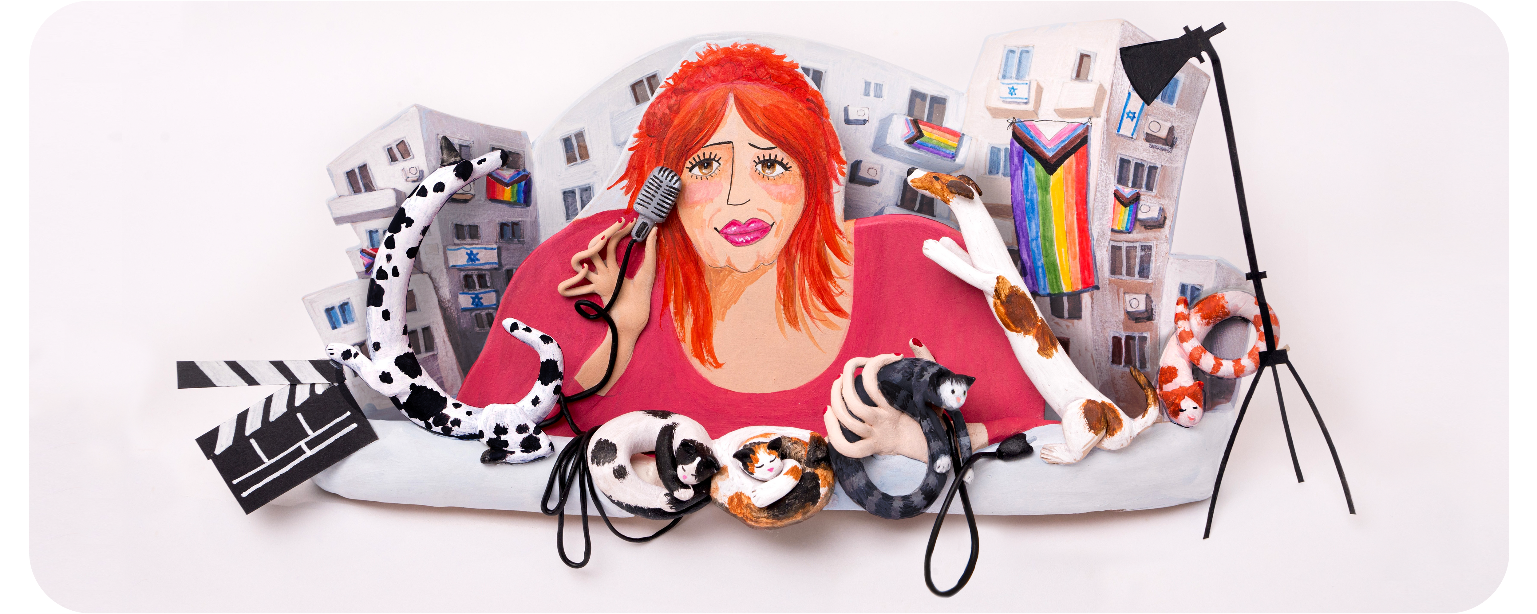 A diorama made of clay. Gila Goldstein is shown in the middle of the diorama from the torso up. She has bright red hair that is cut short with bangs that are swept to each side of her face and is wearing a pink scoop neck, long-sleeve top. She has mascara on and bright pink lipstick and is holding a microphone in her right hand and a black striped cat in her left hand. The GOOGLE logo is spelled out with figures of cats and dogs. There is a black and white film clapper in the bottom left corner and a spotlight tripod on the right. There is a white apartment building behind Gila. Many of the windows have Pride flags hanging from them.