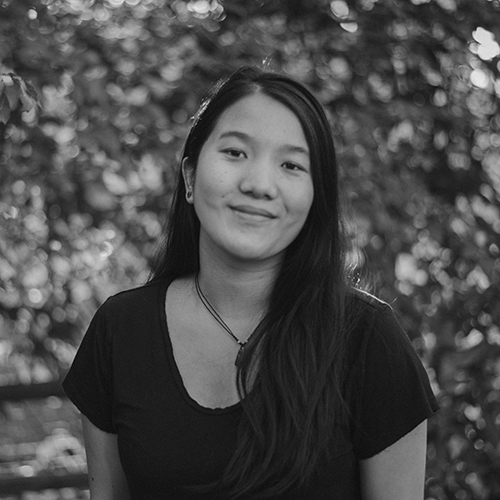Black and white headshot photo of Meeya, standing in front of trees. She has long, straight dark hair with a side part. She is wearing a choker necklace and wearing a scoop neck black t-shirt.