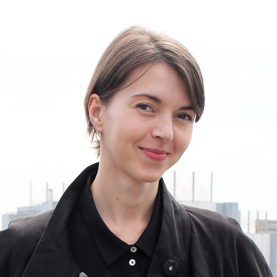 Photo of woman smiling at the camera, standing in front of a skyline. She has short brown hair that is pulled back, fair skin, and dark brown eyes. She is wearing a black collared shirt and black coat. 