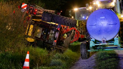 Ein umgekippter Traktor liegt auf der Seite in einem Graben, dahinter sein Anhänger mit Flüssigkeit; Einsatzkräfte vor Ort