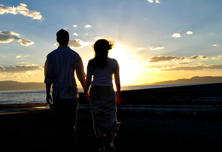 Un hombre y una mujer se toman de la mano en la playa, con su silueta visible frente a la puesta de sol.
