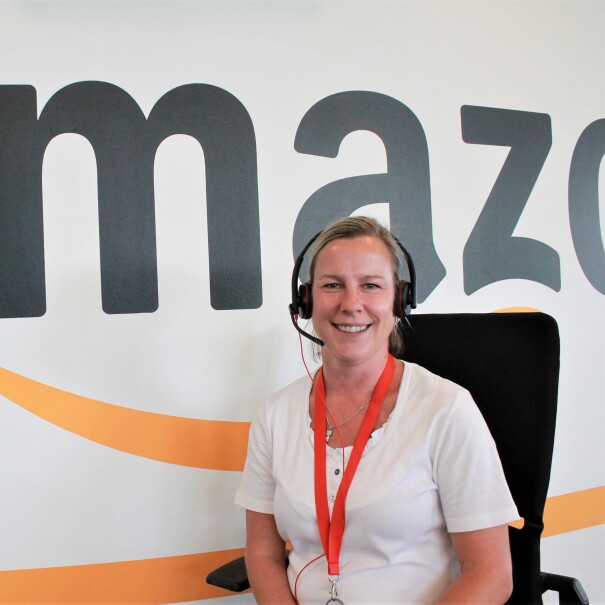 Eine Amazon Kundenservice-Mitarbeiterin mit Kopfhörer vor einer Wand mit Amazon Schriftzug.