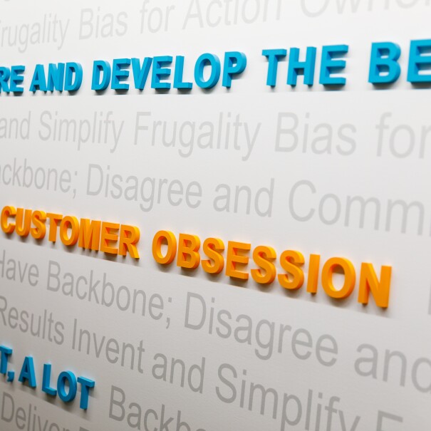 A visual display on a wall at Amazon, highlighting three of the leadership principles, with "customer obsession," "are right, a lot," and "hire and develop the best" in raised letters on a backdrop of other Leadership Principles.