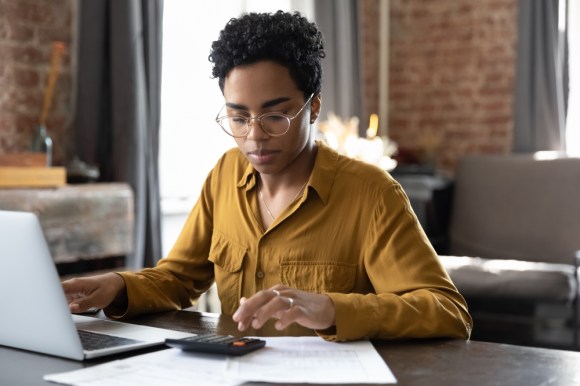 Image of young woman using a calculator and a laptop in home office.