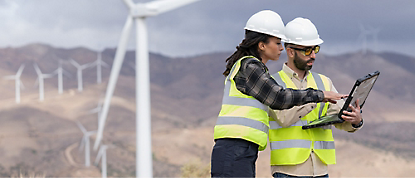 Two construction workers looking at a laptop in front of wind turbines.