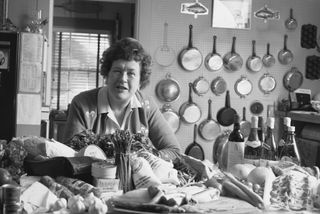 A black and white photo of Julia Childs with an array of fresh ingredients in the kitchen