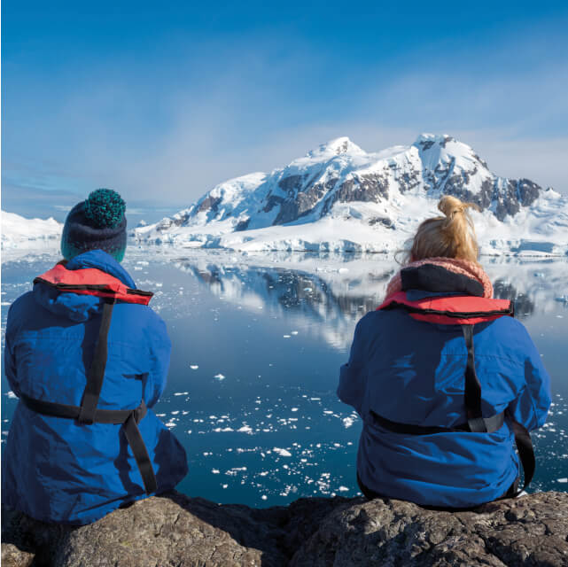 Zwei Menschen mit Rettungswesten schauen mit dem Rücken zur Kamera gedreht auf Eisberge.