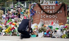 A Black man in a sleeveless T-shirt kneels with his head down in front of dozen of stuffed animals and flowers in front of a brick sign that says Robb Elementary School.