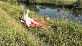 Plage sauvage. Une MILF sexy platine se fait bronzer nue sur la berge de la rivière, un pêcheur regarde au hasard. nue en public. plage naturiste