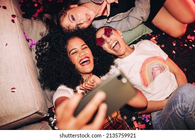 Vibrant selfies for vibrant people. Overhead view of three happy friends taking a selfie while lying on the floor at a house party. Group of cheerful female friends having fun together on the weekend. Stock Photo