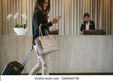 Woman using mobile phone and pulling her suitcase in a hotel lobby. Female business traveler walking in hotel hallway. Stock Photo