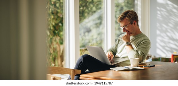 Young man using laptop and smiling at home. Man sitting by table working on laptop computer. Stock Photo