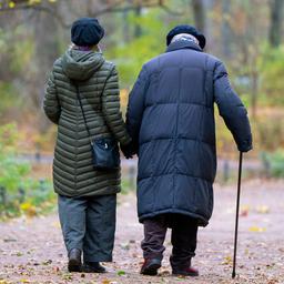 Eine ältere Frau und ein älterer Mann laufen Hand in Hand durch einen herbstlichen Park.