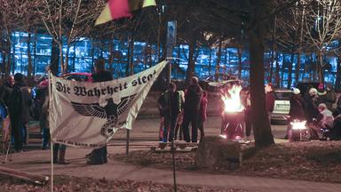 Demonstranten an einer Feuertonne, davor einem Transparent auf dem in Frakturschrift steht "die Wahrheit siegt", darunter ist ein Symbol ähnlich einem Reichsadler zu erkennen.