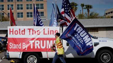 Ein Mann läuft mit einer US-amerikanischen Flagge vor einem mit Wahlplakaten versehenen Truck vorbei. (Archivbild vom 14.12.2020)