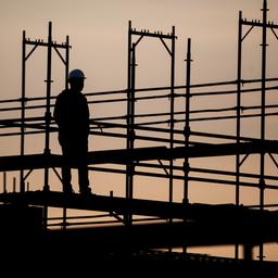 Die Silhouette eines Bauarbeiters zeichnet sich auf einer Baustelle vor dem verfärbten Morgenhimmel ab.