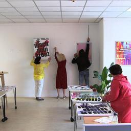 Vier Menschen stehen in einem Raum und hängen Plakate für eine Kunstausstellung auf.