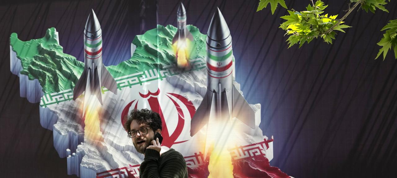 Ein Passant, der mit einem Handy telefoniert, geht in Teheran an einem Banner vorbei, das den Abschuss von Raketen von der iranischen Landkarte zeigt. 