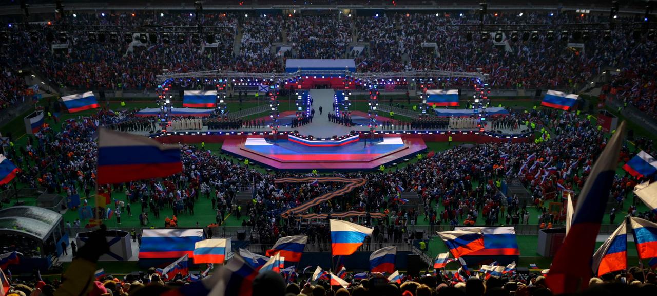 Menschen schwenken russische Fahnen, als Wladimir Putin die Bühne betritt, um eine Rede während eines patriotischen Konzerts zu halten.