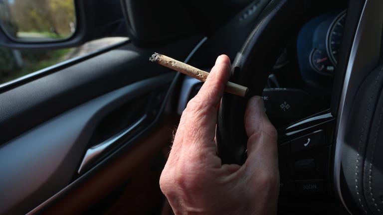 Ein Mann sitzt mit einem Joint zwischen den Fingern am Steuer eines Autos