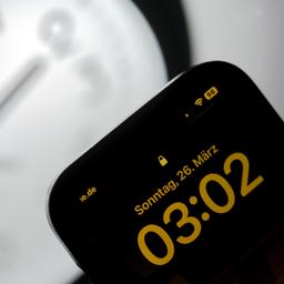 Ein Smartphone zeigt die Uhrzeit "03:03" an, während auf einer analogen Uhr noch 02:03 Uhr ist.