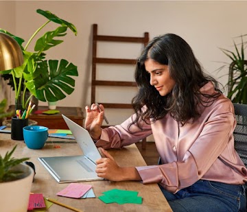 A person with dark hair sits at a desk holding an inverted Chromebook in their left hand and a stylus in their right hand.