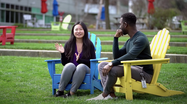 In a grassy outdoor setting, two people sit across from each other, smiling and engaged in conversation