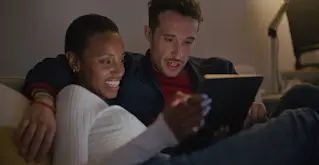 Um casal num sofá vê conteúdo multimédia no seu tablet.