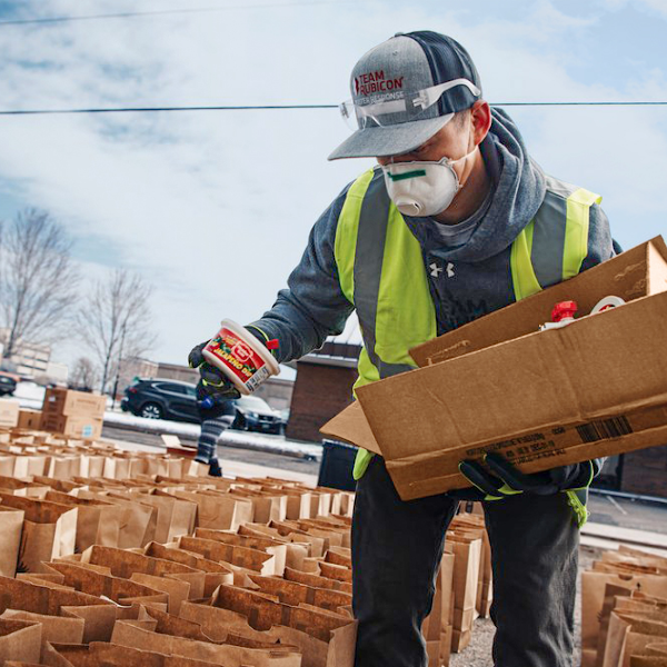 A first responder wearing a face mask and yellow safety vest helps organize food and supplies into paper bags