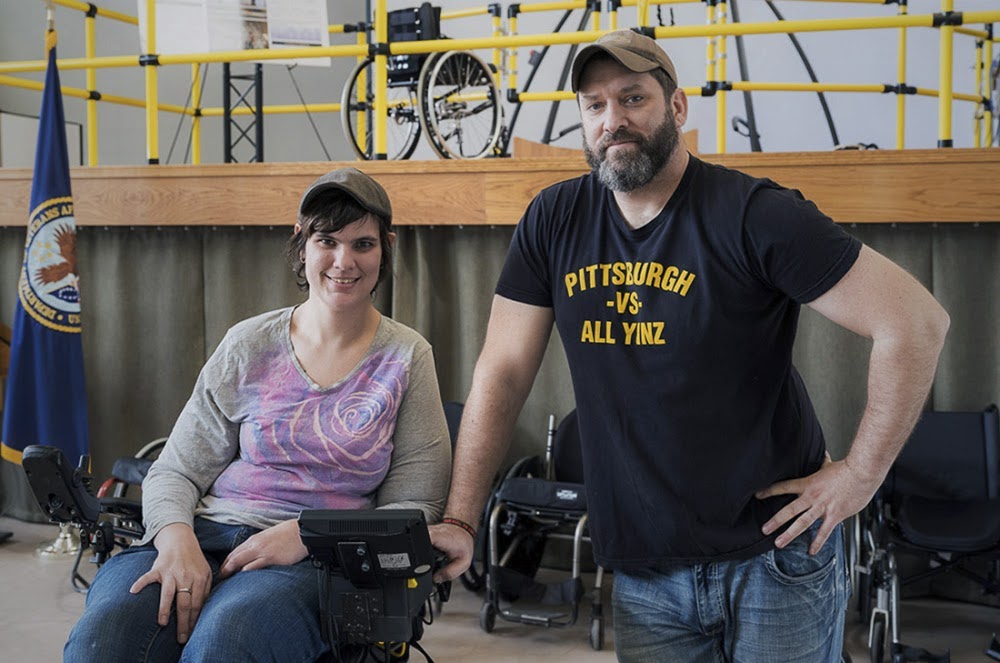 A person in a wheelchair next to a person standing with their hand resting on the arm of the wheelchair.