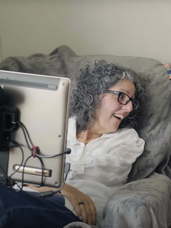 A woman with grey hair and black glasses sits in a chair with a screen in front of her looks to the side smiling