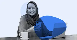 Uma mulher com um hijabe sorri enquanto usa o seu smartphone e portátil.