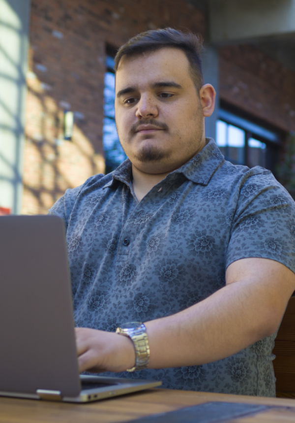 A man wearing a blue patterned button-down shirt sits and types on a laptop