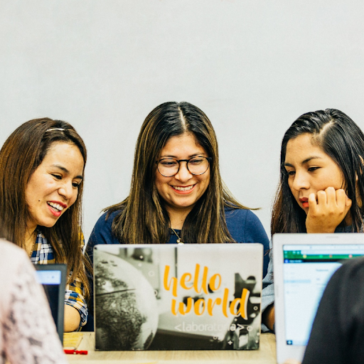 Three young women sit together at desk and look at laptop