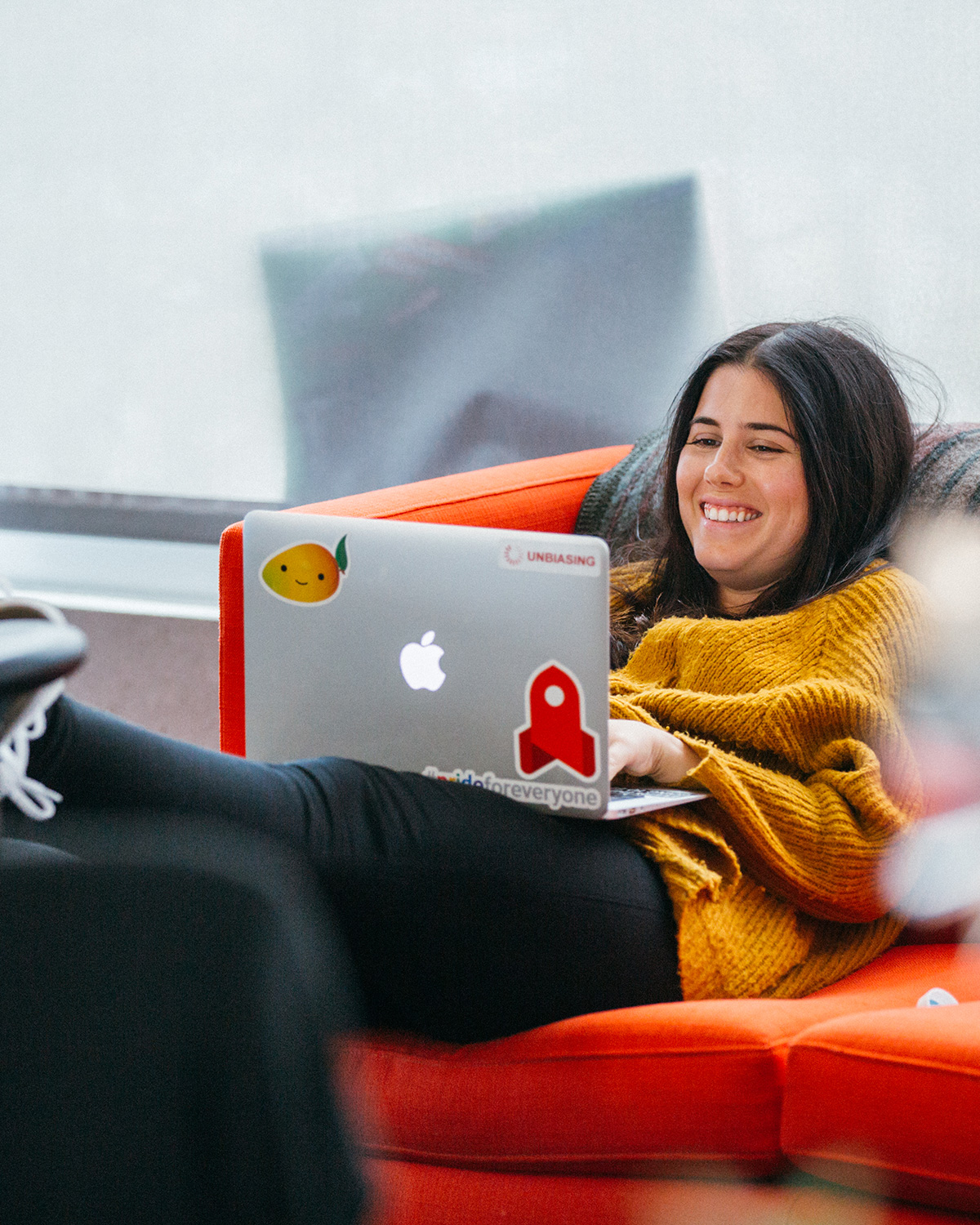A woman with her feet up, in an orange chair, smiling while she works on her laptop.