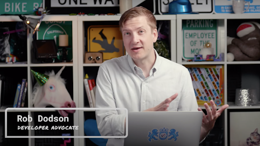 A man in a dress shirt sits behind a laptop in front of a bookcase with collectibles