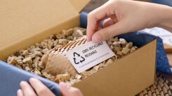 A shipping box filled with reusable packing material, with a hand visible holing a small card that reads "Reusable, Recyclable" 