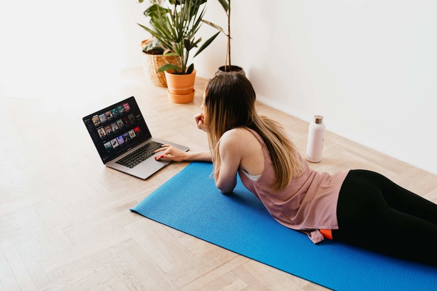 A young woman lies on a yoga mat while looking on her laptop.