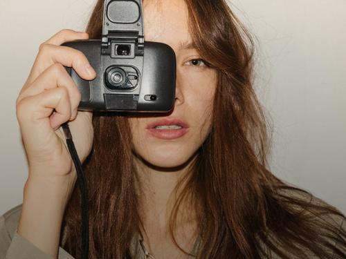 Eine Frau mit braunen Haaren, die eine alte Kamera vor einer grauen Wand benutzt und ein olivfarbenes Oberteil trägt.