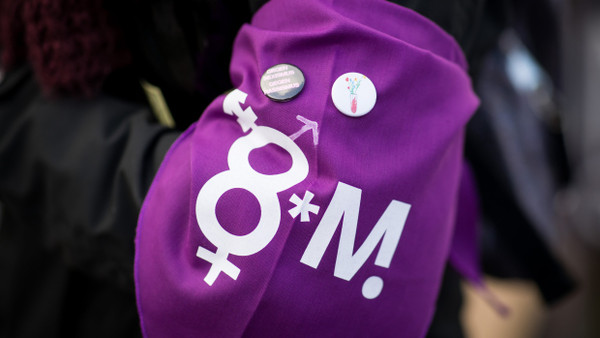Eine Frau trägt ein lila Tuch mit verschiedenen Gendersymbolen und Buttons an ihrem Arm bei einer Kundgebung zum Internationalen Frauentag 2019 vor dem Hamburger Rathaus.