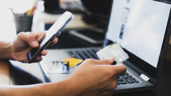 Ein Mann verwendet zum Onlineshopping eine Kreditkarte, ein Smartphone und einen Laptop.