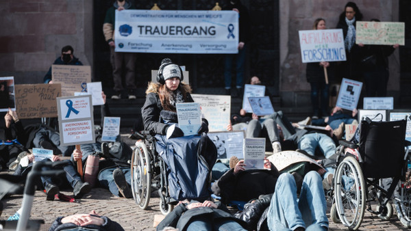 Im Sitzen und Liegen: Eine Erkrankte demonstriert im Rollstuhl, Angehörige und Freunde legen sich vor dem Frankfurter Rathaus Römer auf den Boden - stellvertretend für die zu Hause liegenden ME/CFS-Patienten.