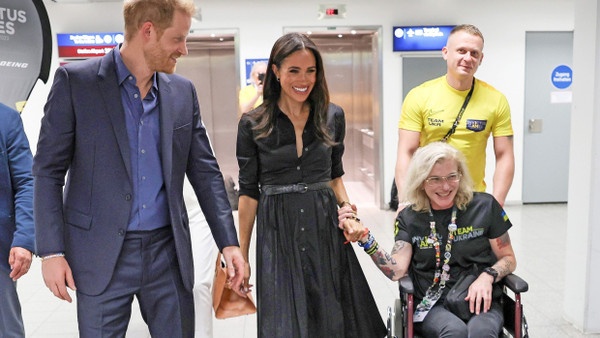 Prinz Harry, seine Frau Meghan und eine Teilnehmerin der Invictus Games halten sich an den Händen. Ein Mann schiebt den Rollstuhl der Teilnehmerin.