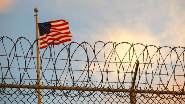 Eine US-amerikanische Flagge weht hinter einem Stacheldrahtzaun im Wind. (Symbolbild)