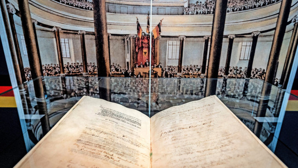 Unscheinbar, aber bedeutend: Die Verfassungsurkunde vom 28. März 1849, in der Paulskirche vor einem zeitgenössischen Bild der Nationalversammlung präsentiert.