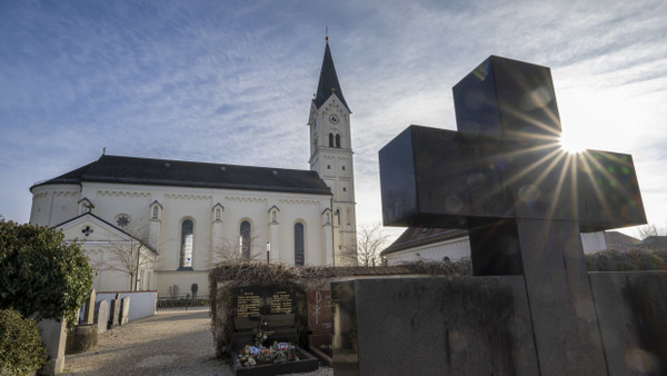 Das Kreuz eines Grabsteins ragt vor der katholische Kirche St. Nikolaus in Garching an der Alz in den Himmel.