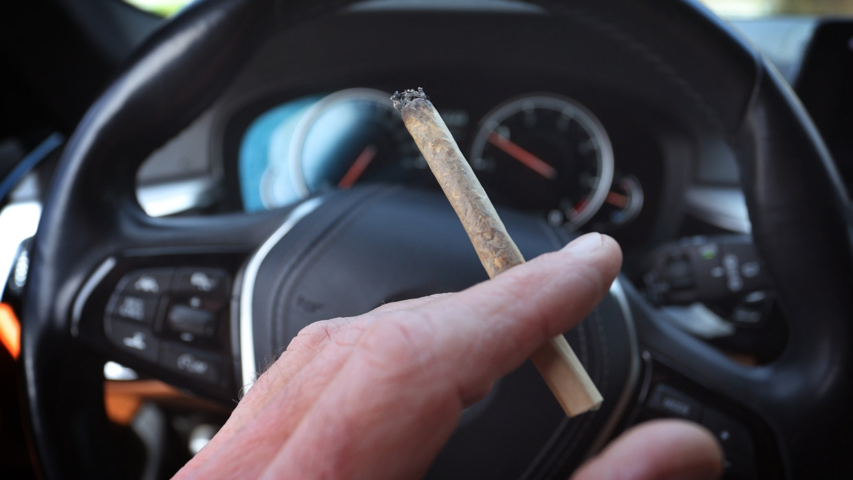 Ein Mann sitzt mit einem Joint zwischen den Fingern am Steuer eines Autos. (Symbolbild)